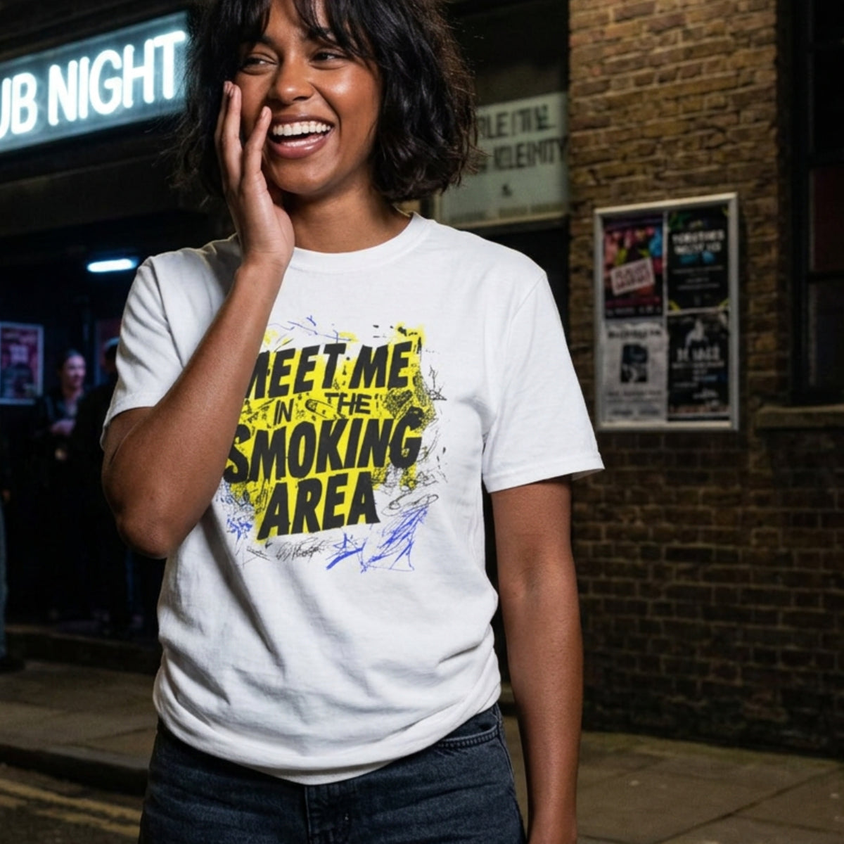 A woman smiling on a city street at night wearing a white graphic t-shirt with yellow and blue text that reads 'MEET ME IN THE SMOKING AREA'.