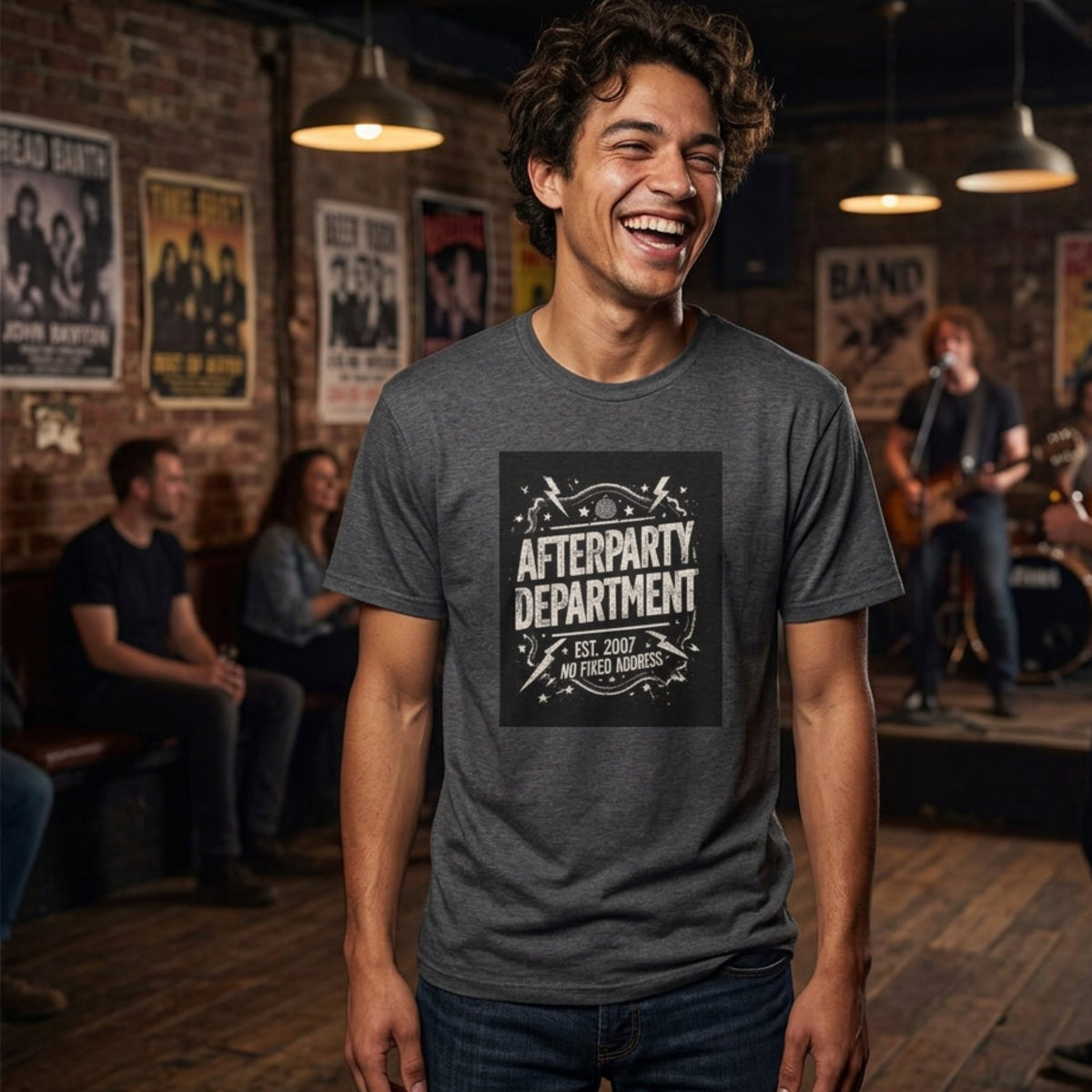 A man wearing a dark gray heather t-shirt with a black graphic that reads 'AFTERPARTY DEPARTMENT EST. 2007 NO FIXED ADDRESS' in a band club setting with a brick wall and band members in the background.