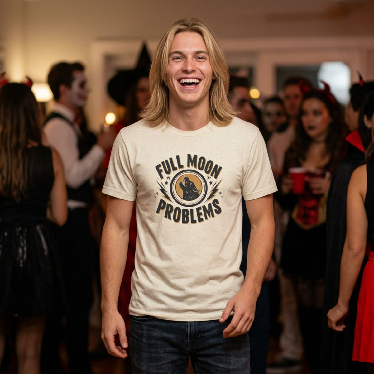 A man with long blonde hair is smiling and laughing in a crowded indoor party setting, wearing a cream-colored graphic t-shirt with the text 'FULL MOON PROBLEMS' and a crescent moon graphic.