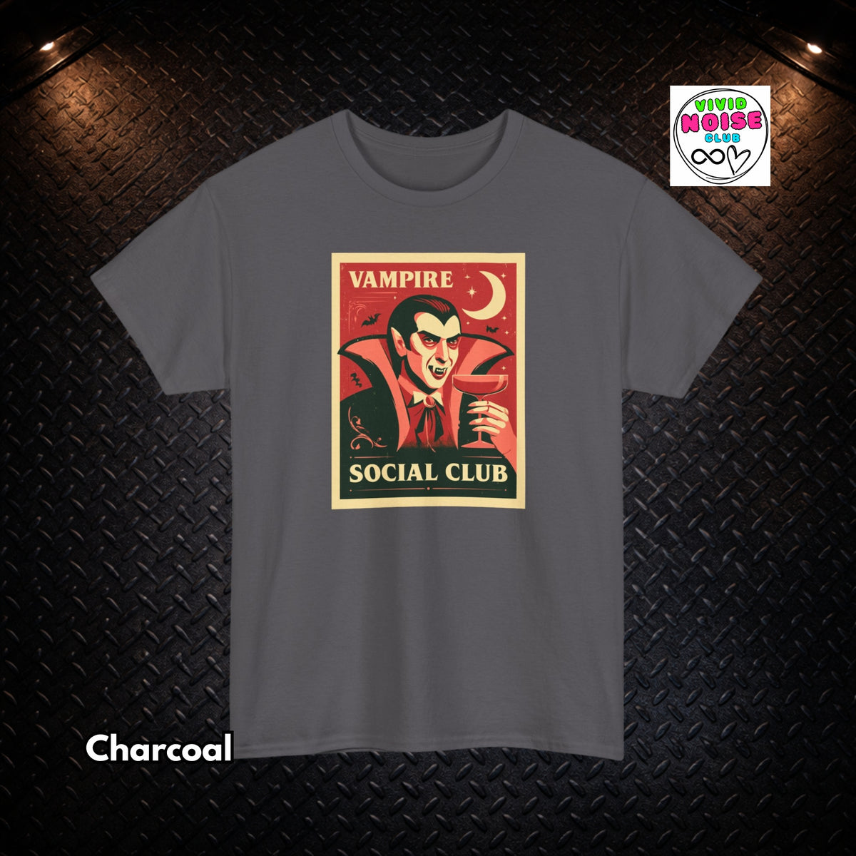 A charcoal gray short-sleeve t-shirt with a vintage-style graphic print of a vampire holding a wine glass, with the text 'VAMPIRE SOCIAL CLUB'. The t-shirt is displayed flat against a dark textured diamond plate background with dramatic overhead lighting.