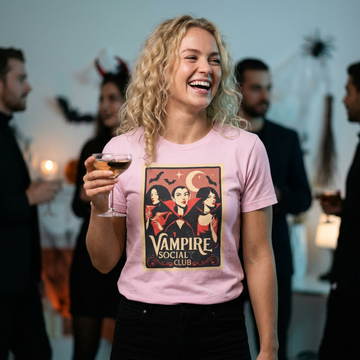 A woman at a Halloween party wearing a light pink graphic t-shirt with a 'Vampire Social Club' illustration. She is holding a glass of wine and smiling, with blurred partygoers and spooky decorations in the background.