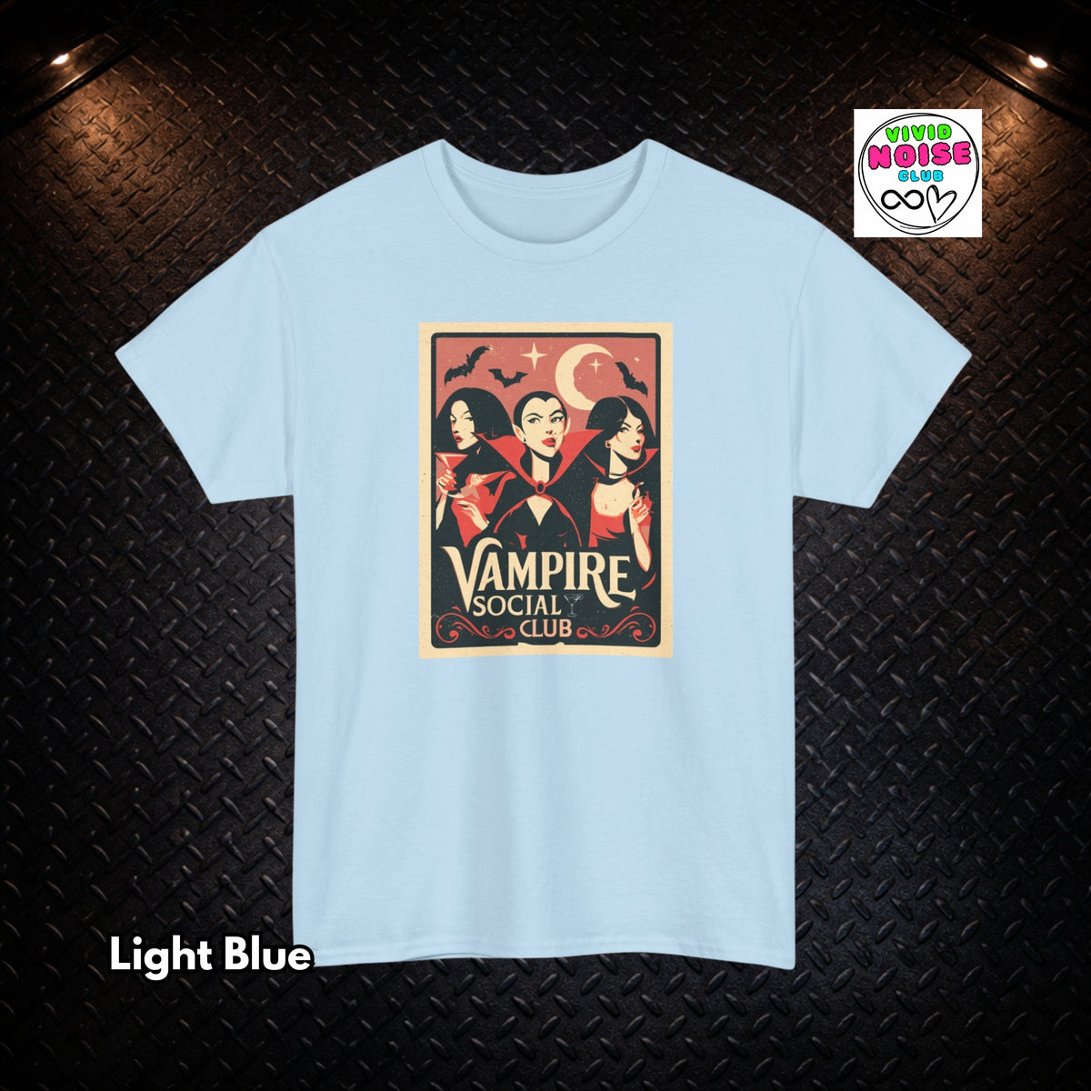 A light blue short-sleeve t-shirt with a vintage-style graphic print of three vampires under a crescent moon with the text 'Vampire Social Club'. The t-shirt is displayed flat against a dark textured background with 'Light Blue' text in the bottom-left corner and a 'Vivid Noise Club' logo in the top-right corner.