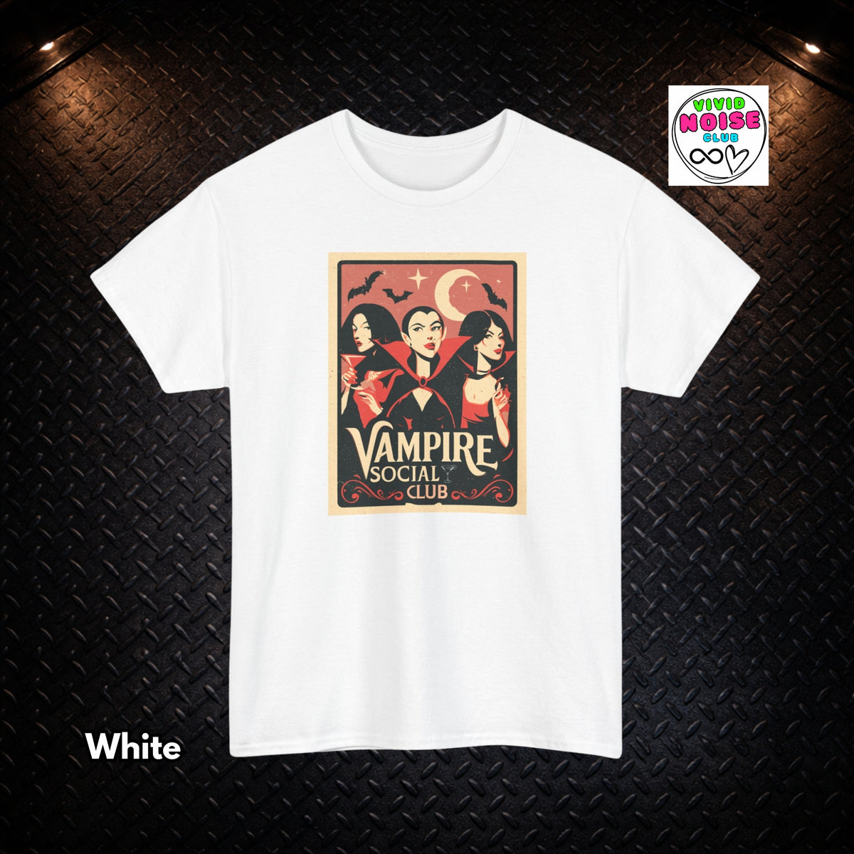 A white short-sleeve crewneck t-shirt with a vintage-style graphic print of three vampires under a crescent moon and stars, labeled 'Vampire Social Club'. The t-shirt is displayed flat against a black textured diamond plate background with warm overhead lights.