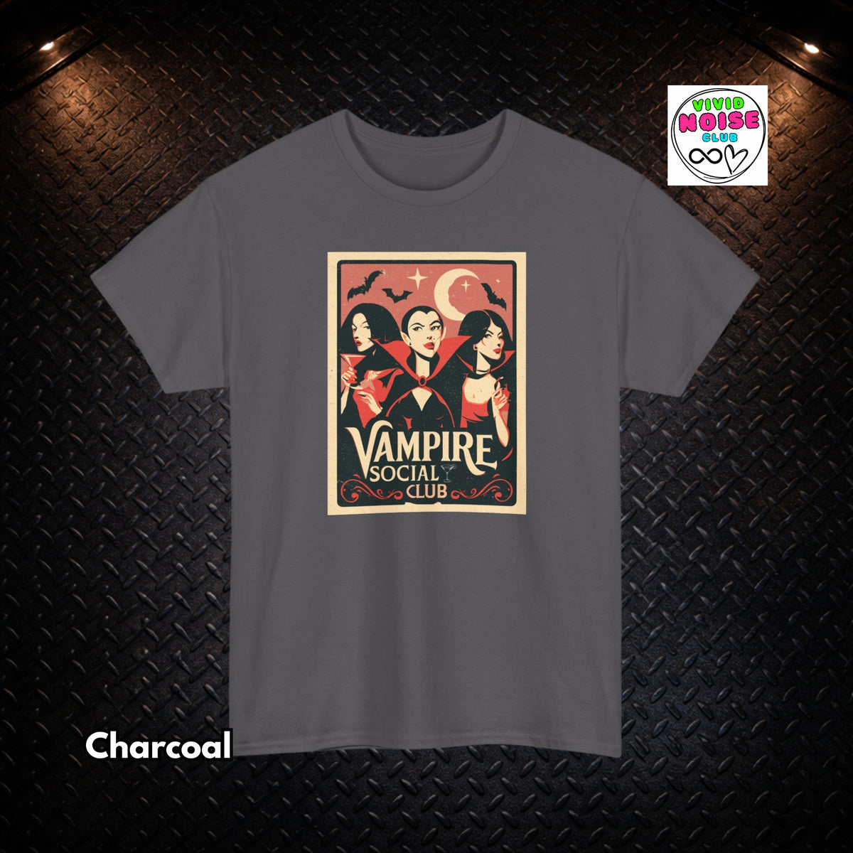 A charcoal gray t-shirt with a vintage-style graphic print of three vampires under a crescent moon and stars, with the text 'The Vampire Social Club'. The t-shirt is displayed flat against a dark textured diamond plate background with dramatic overhead lighting.
