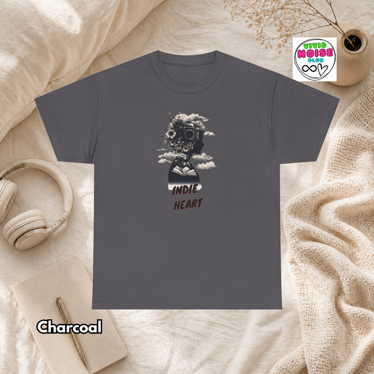 A charcoal gray t-shirt laid flat on a beige fabric background, featuring a graphic of a gas mask surrounded by flowers and the text 'INDIE HEART'. The shirt is styled with a pair of headphones, a notebook, a small plant, and a cozy blanket, with a 'Charcoal' color label and a 'Vivid Noise Club' logo in the corners.