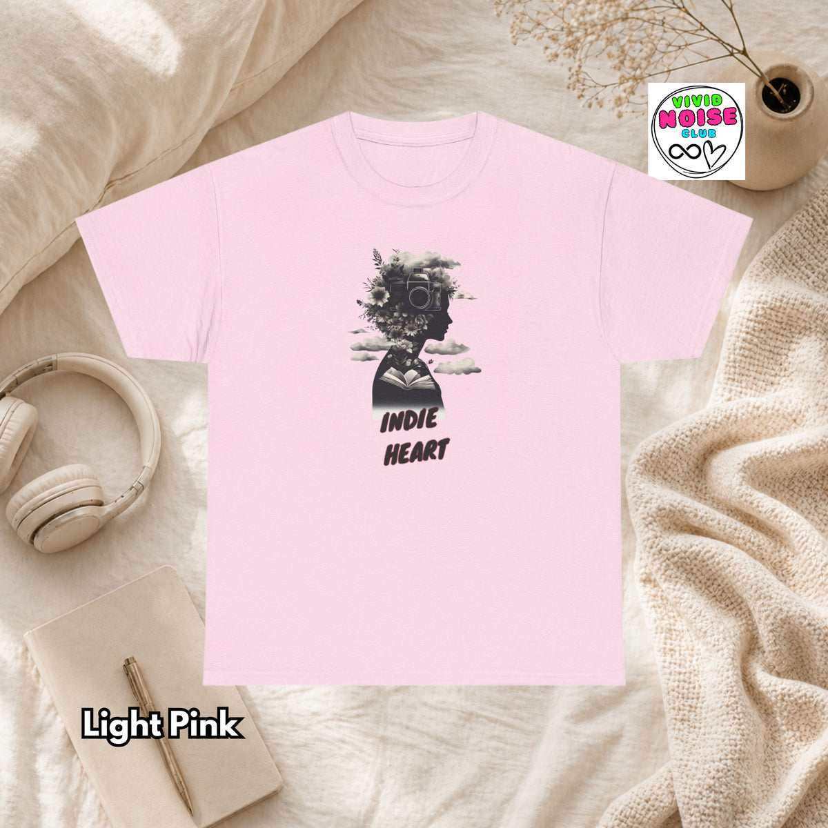 A light pink short-sleeve t-shirt laid flat on a soft beige fabric background, featuring a black graphic of a silhouette with a camera and the words 'INDIE HEART'. A 'Vivid Noise Club'' brand sticker is visible in the top right, and 'Light Pink' is written in the bottom left.