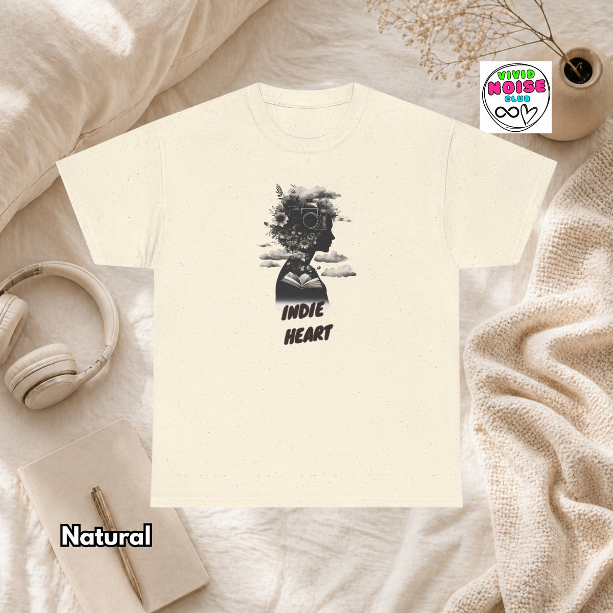 A cream-colored t-shirt with a black graphic of a camera and the text 'INDIE HEART' is displayed in a cozy flat-lay setting with a knit blanket, headphones, and a notebook. A brand logo for 'Vivid Noise Club' is visible in the top right, and the word 'Natural' is written in the bottom left.