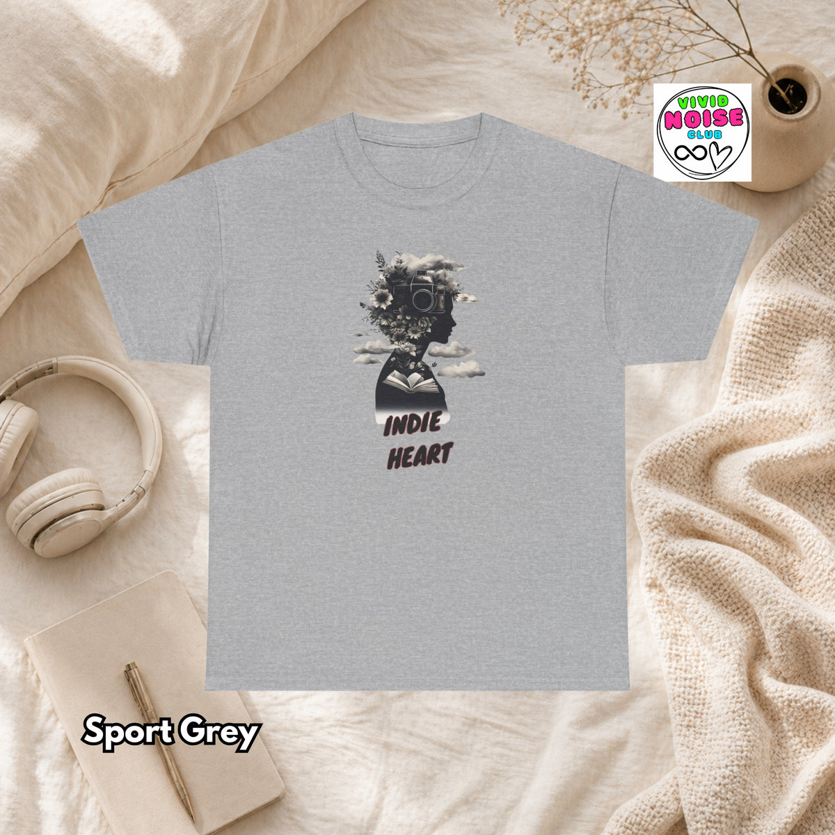A light grey heather t-shirt laid flat on a cozy beige bed with a graphic of a silhouette and flowers with the text 'INDIE HEART'. The image includes a 'Vivid Noise Club'' brand logo in the top right and a 'Sport Grey' text label in the bottom left, surrounded by lifestyle props like headphones, a notebook, and a blanket.
