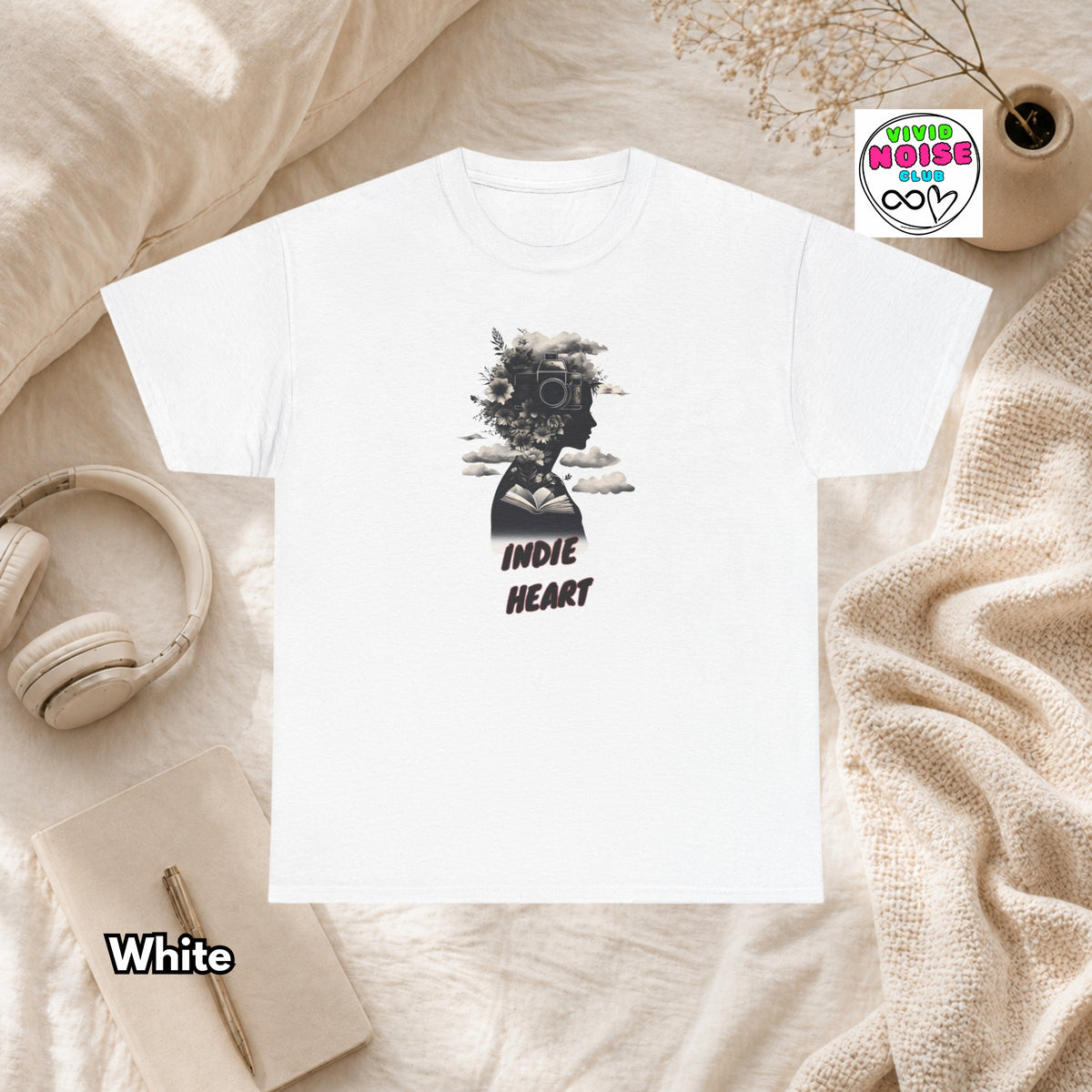 A white short-sleeve T-shirt laid flat on a soft beige fabric surface, featuring a black graphic of a face with headphones and the words 'INDIE HEART' below it. A small 'Vivid Noise Club' logo appears near the top right, and the word 'White' appears near the bottom left.