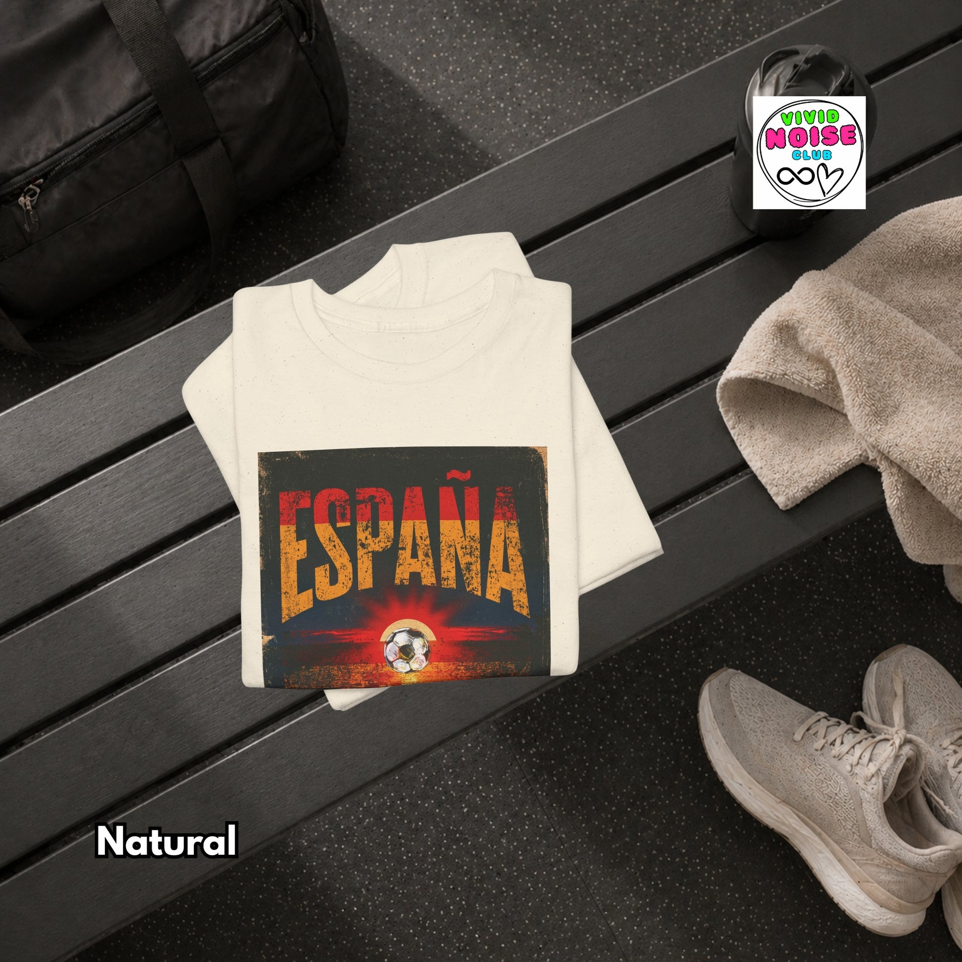 A folded white t-shirt with a distressed graphic reading 'ESPAÑA' in orange and yellow over a dark background, styled on a dark bench with a towel, shoes, and a bag nearby. 