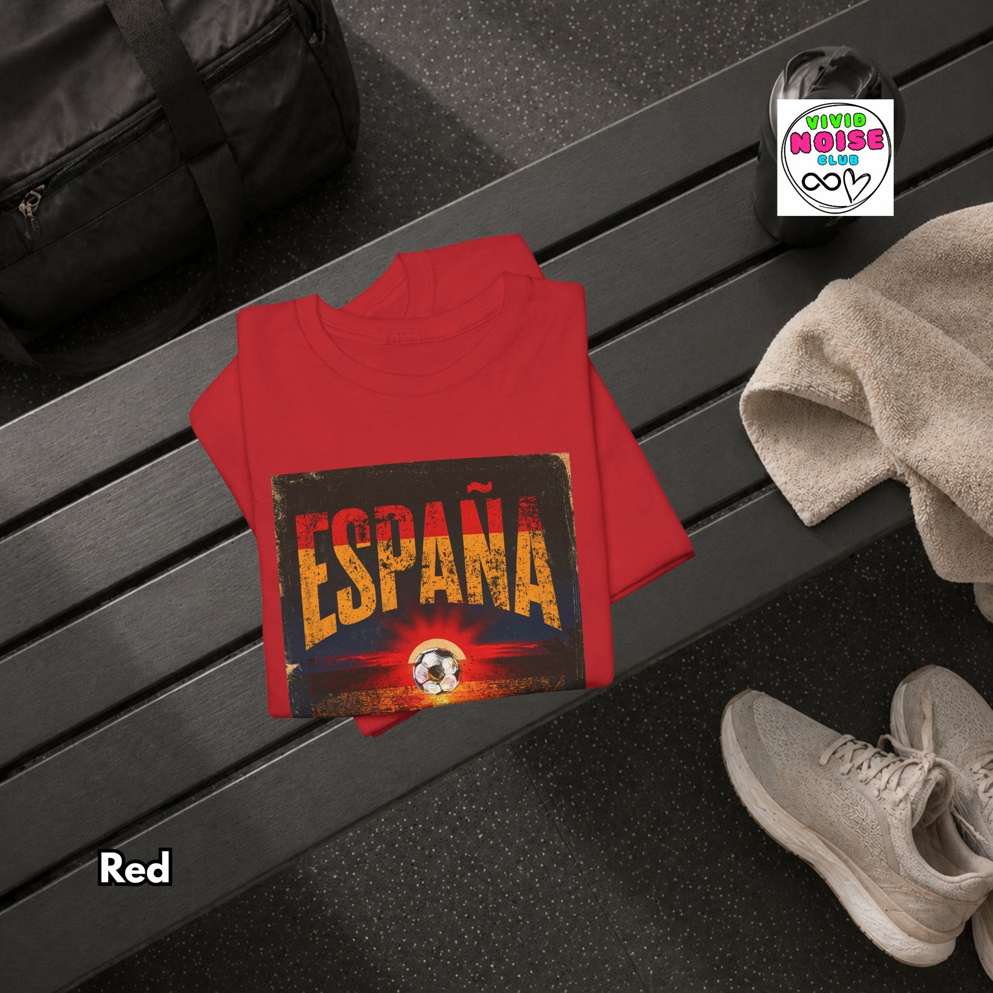 A red folded T-shirt is placed on a dark wooden bench in a gym setting, with a black duffel bag, towel, and gray sneakers nearby. The shirt features a distressed graphic that reads 'ESPAÑA' in large yellow letters over a sunset-style design with a soccer ball at the bottom.