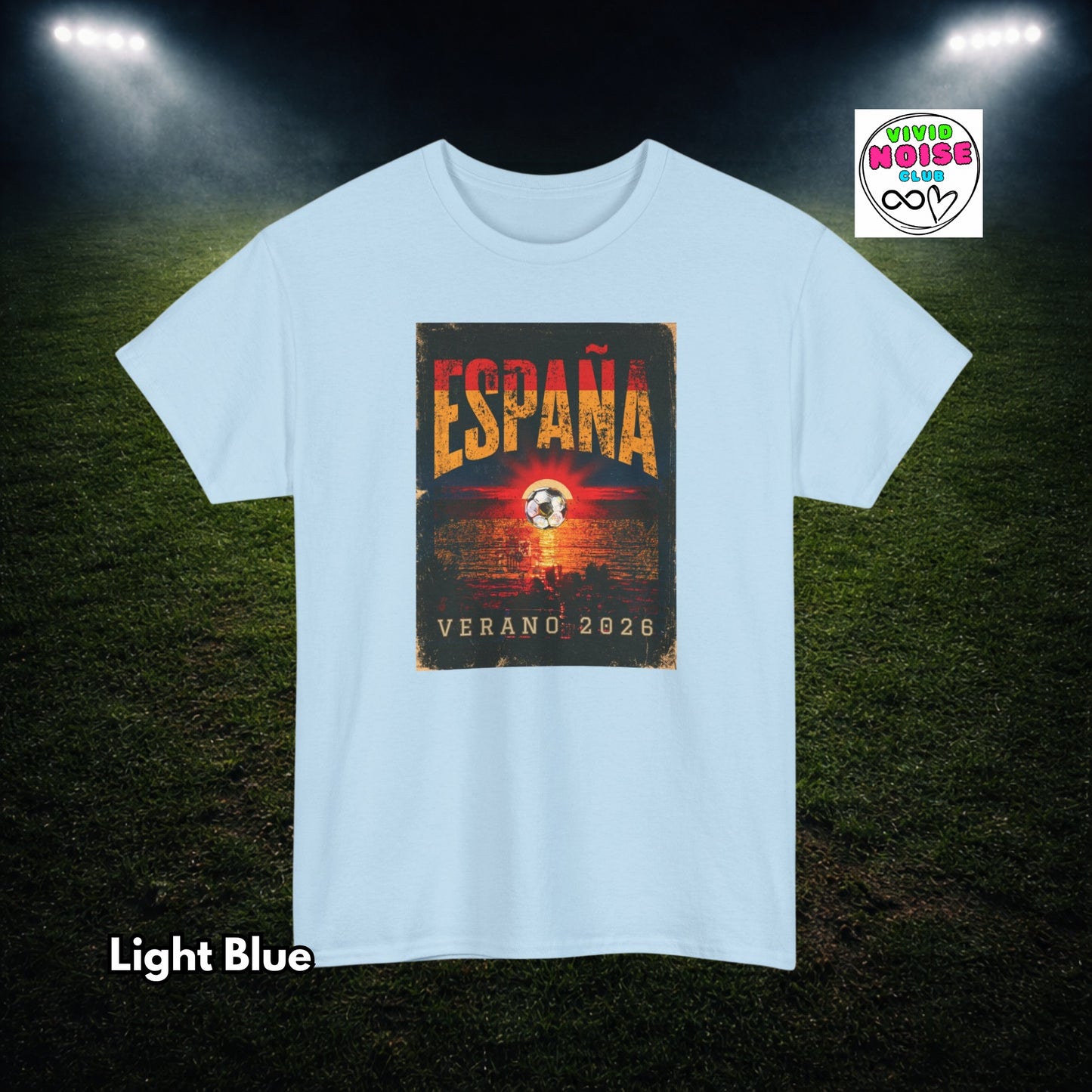 A light blue short-sleeve t-shirt with a rectangular graphic on the chest reading 'ESPAÑA VERANO 2026' in orange and yellow tones. The shirt is displayed against a dark grassy field background with stadium lights shining down.