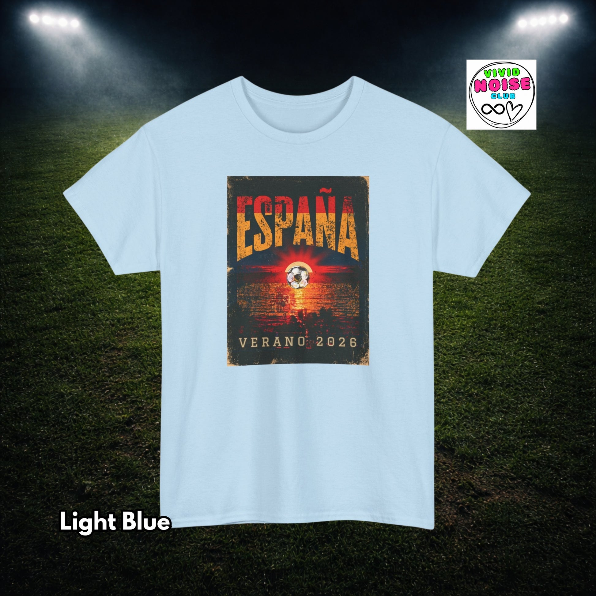 A light blue short-sleeve t-shirt with a rectangular graphic on the chest reading 'ESPAÑA VERANO 2026' in orange and yellow tones. The shirt is displayed against a dark grassy field background with stadium lights shining down.