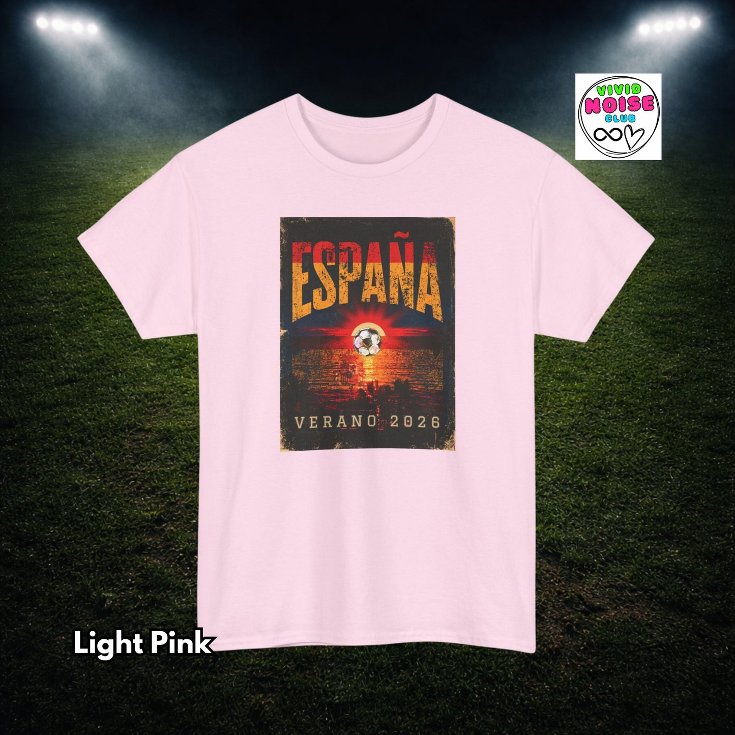 A light pink short-sleeve t-shirt with a central graphic print featuring the word 'ESPAÑA' in distressed orange letters over a dark background with a soccer ball motif and the text 'VERANO 2026'. The shirt is displayed against a dark grass field background with stadium lights, and a 'Vivid Noise Club' brand logo is visible in the top right corner.